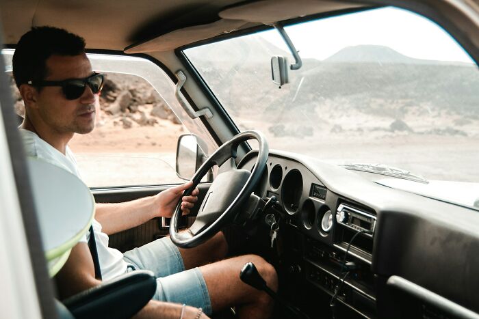 Truck driver wearing sunglasses behind the wheel, cruising through a remote desert landscape with mountains in the distance.