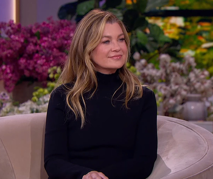 Ellen Pompeo sitting on a couch, wearing a black top, with floral decorations in the background.