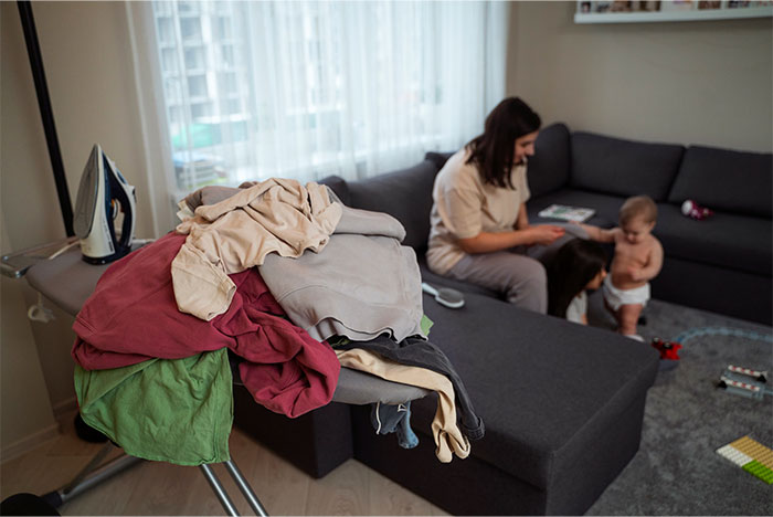 Pile of messy clothes on an ironing board in a living room while a woman and children play in the background