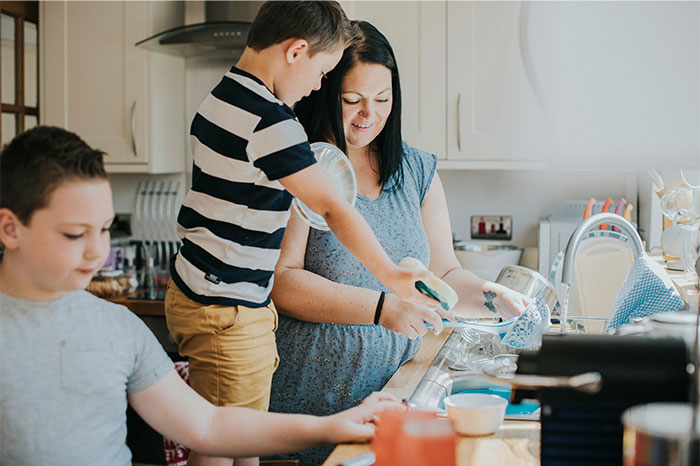 Woman and children washing dishes together in kitchen, highlighting family effort amid messy home cleanup challenges.