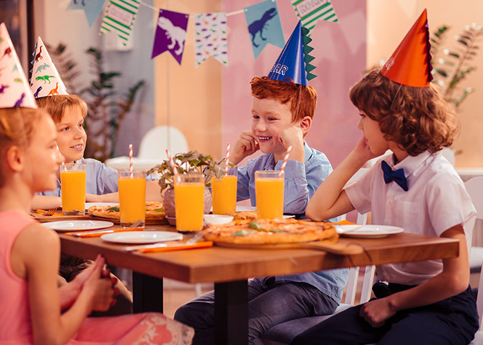 Kids wearing party hats sitting around a table with pizza and juice at a birthday party, capturing a kid&rsquo;s party scene.