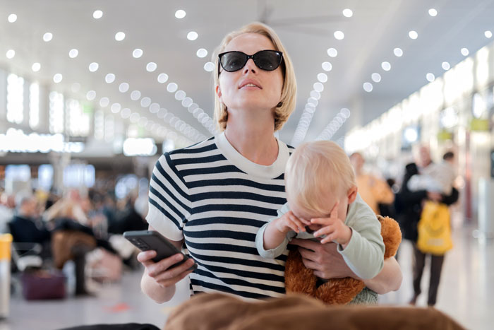 Woman in sunglasses holding a child and phone at airport, showing entitled mother insisting on paying upgrade.