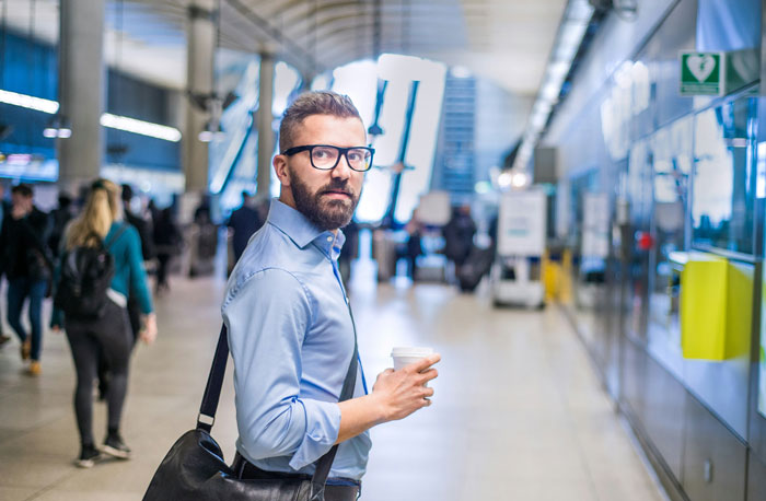 Man with glasses and beard holding coffee, standing in busy terminal, highlighting entitled mother insists paying upgrade.