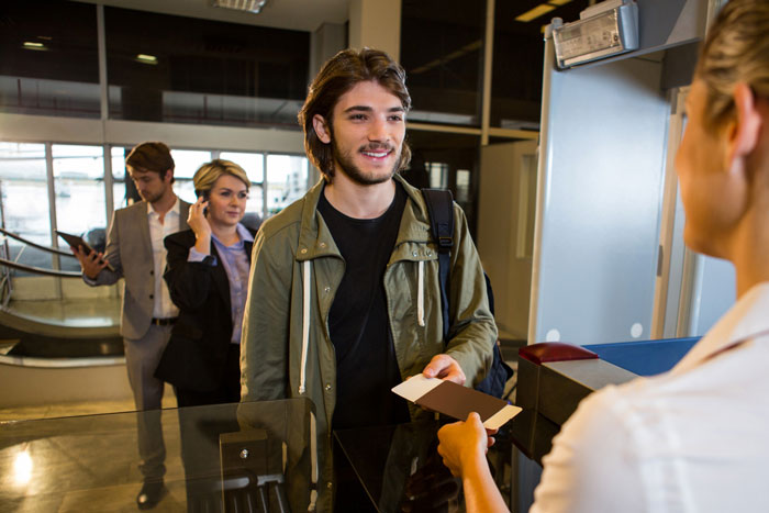 Young man at airport check-in counter handing over documents as entitled mother insists on paying for upgrade behind him.