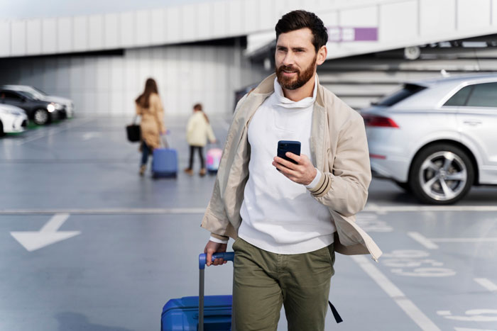 Man with suitcase holding phone in parking lot, illustrating entitled mother insists paying upgrade travel concept