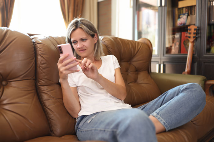 Stepmom sitting on couch looking upset and frustrated while using a smartphone in a cozy living room setting