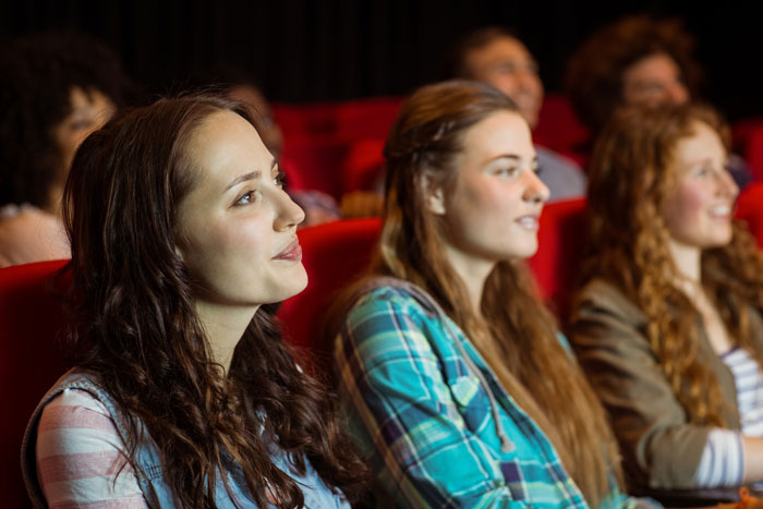 Young women seated in a theater, watching a drama about an ex and narcissist causing conflict in relationships.