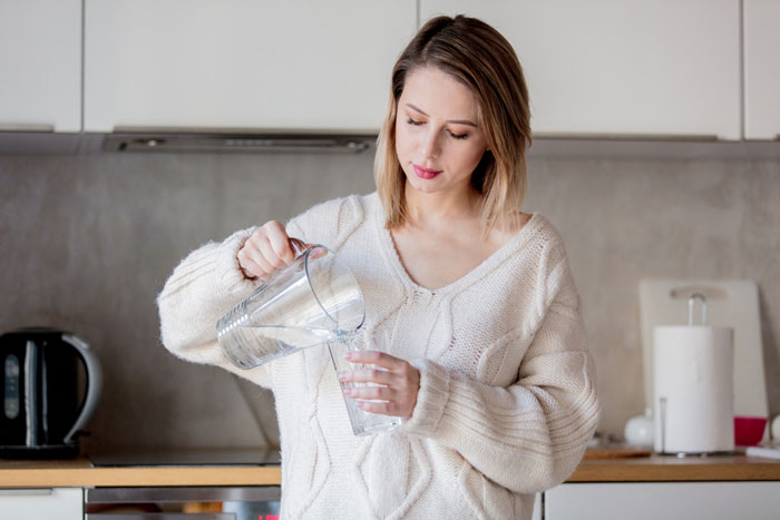 Woman in a cozy sweater pouring water into a glass in a kitchen, highlighting risks of prolonged fasting effects.
