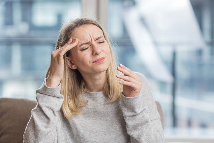 Woman in a gray sweater showing discomfort and headache, illustrating risks of prolonged fasting in a modern indoor setting.