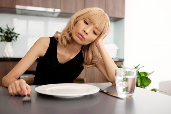 Young woman looking tired and weak while sitting at a table with an empty plate, illustrating prolonged fasting dangers.