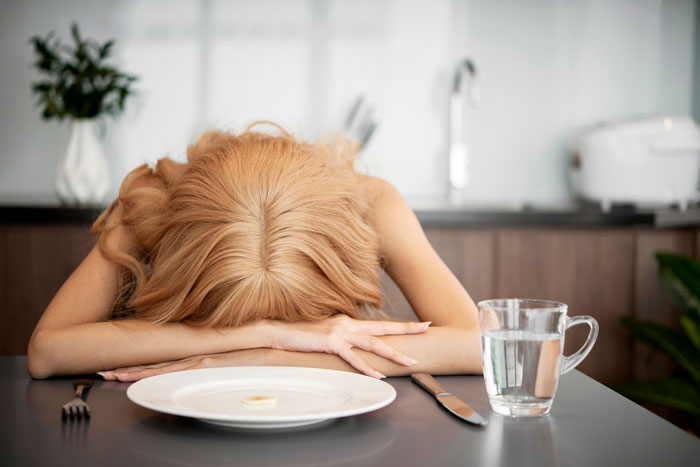 Woman resting head on table with a small piece of food on plate and glass of water, illustrating prolonged fasting risks.