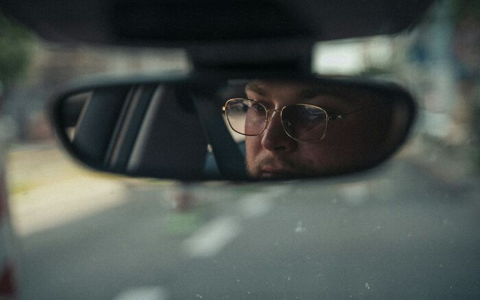 Close-up of a man wearing glasses reflected in a car rearview mirror, symbolizing moments people realized their jerk friend.