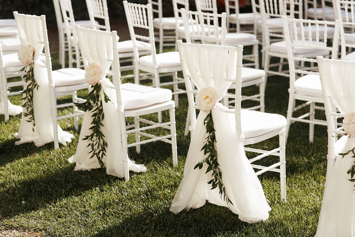 White wedding chairs decorated with flowers and greenery set up outdoors for a ring bearer and guest seating arrangement.