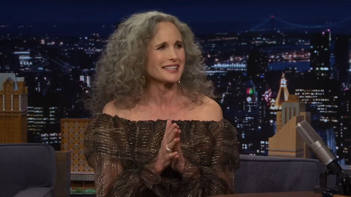 Actress with curly gray hair wearing a brown off-shoulder dress, discussing Hollywood hypocrisy about aging on a talk show.