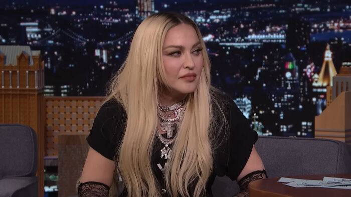 Female celebrity with long blonde hair wearing layered necklaces and a black outfit on a nighttime talk show set.