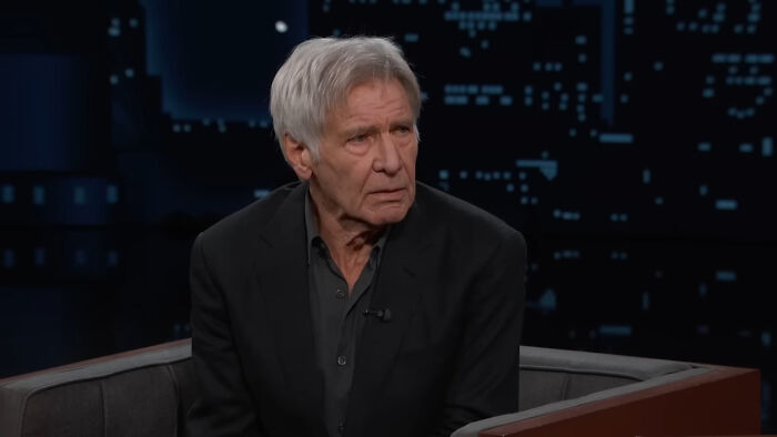 Older male celebrity in a dark suit on a talk show highlighting Hollywood hypocrisy about women aging versus men.