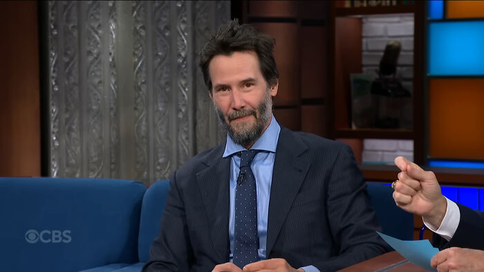 Keanu Reeves on a talk show set, wearing a suit and tie, illustrating Hollywood hypocrisy on aging differences.