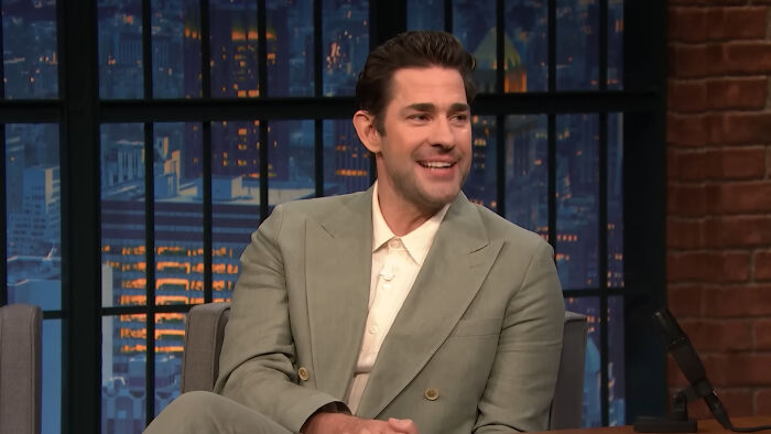 Actor smiling in a grey suit on a talk show set illustrating Hollywood hypocrisy about men aging like fine wine.