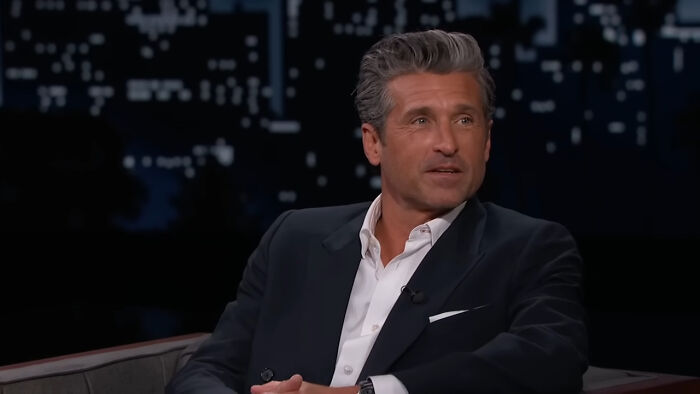Actor in a dark suit on a late night talk show set, highlighting Hollywood hypocrisy about men aging like fine wine.