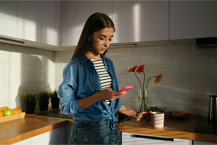 Woman in kitchen holding a pink phone and cup, reflecting on a man skipping his wife&rsquo;s first Mother&rsquo;s Day celebration.
