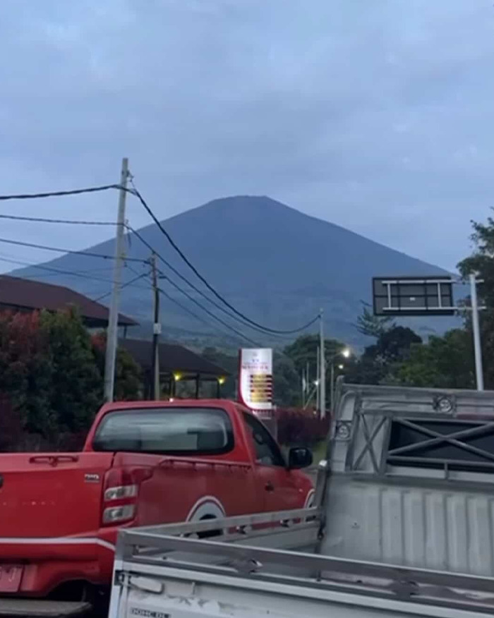 Red pickup trucks parked on street with active volcano visible in the background on a cloudy day.