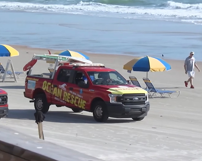 Red ocean rescue truck parked on a beach near umbrellas and lounge chairs by the ocean shore.
