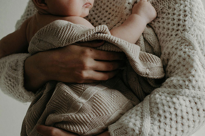 Teen girl holding infant wrapped in beige blanket, showing signs of sleep deprivation and stress from daily babysitting.