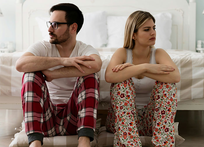 Married couple sitting apart on bedroom floor, showing tension over having more kids and relationship struggles. Married couple sitting apart on bedroom floor, showing tension over having more kids and relationship struggles.
