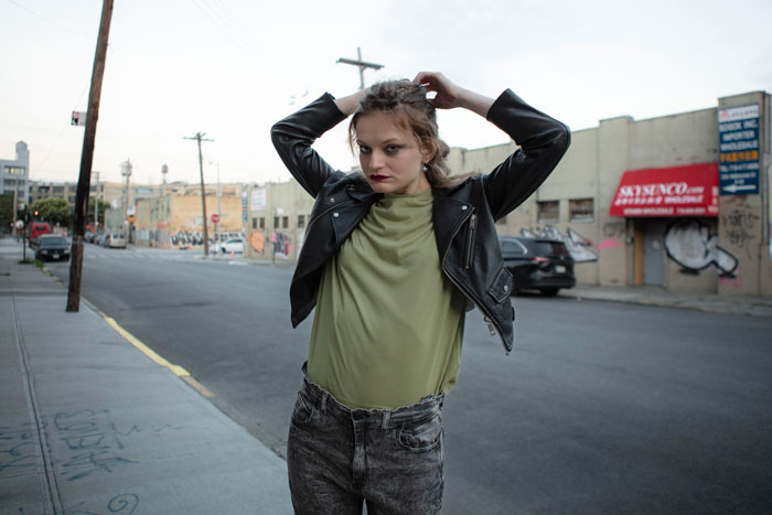 Young woman adjusting her hair on an urban street, wearing a leather jacket and casual clothes near a friend stepdaughter pool party setting.