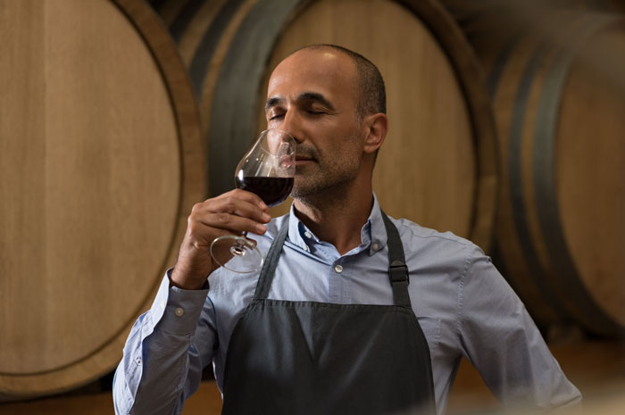 Man in apron sampling red wine in cellar with large barrels, enjoying aroma with eyes closed and relaxed expression.