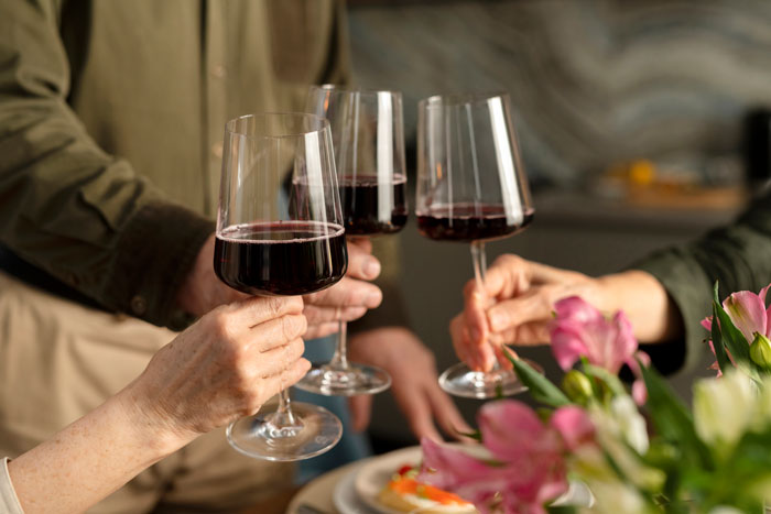 Three friends holding red wine glasses together for a sample toast during a casual gathering with flowers nearby.