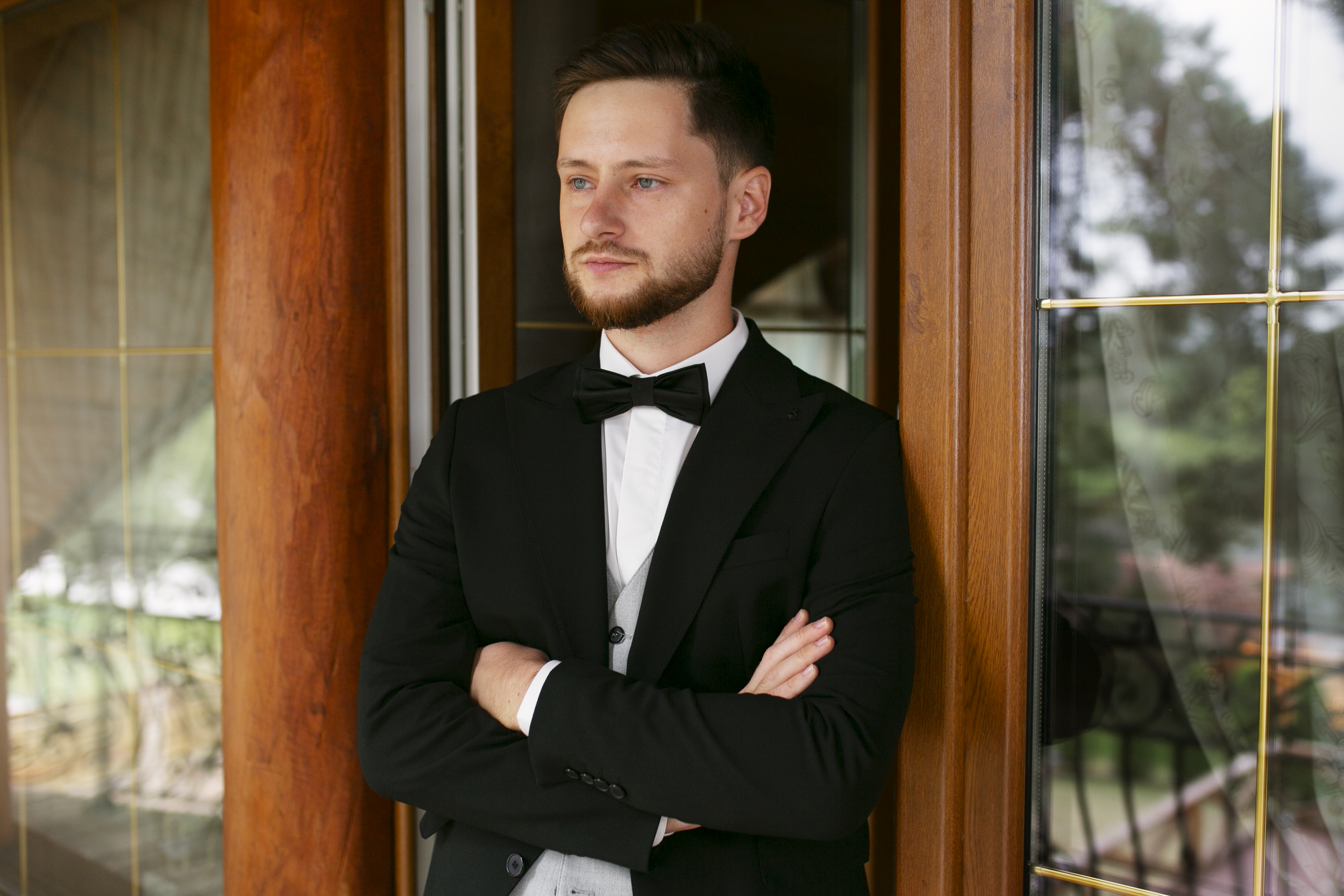 Best man in formal black suit and bow tie standing with arms crossed by a wooden door in wedding setting.