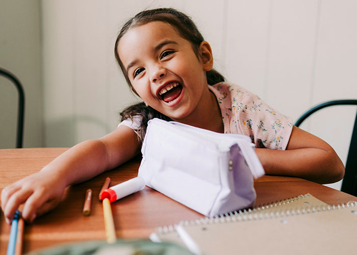 Smiling young girl at a table with school supplies, capturing the funniest and most inappropriate things kids say.