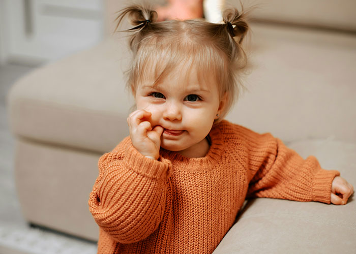 Toddler with pigtails wearing an orange sweater, displaying a playful expression amid funny and inappropriate kids' blurts shared by parents.