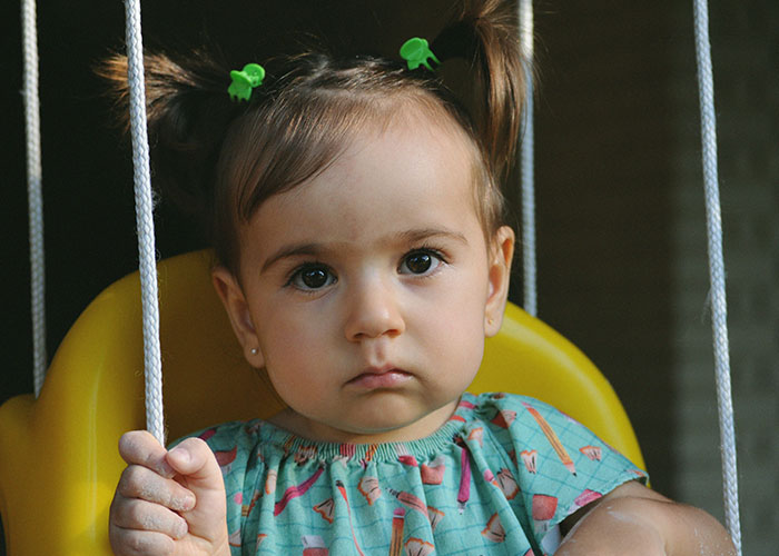 Toddler with pigtails on yellow swing, looking serious, representing funniest and most inappropriate kids blurts shared by parents.