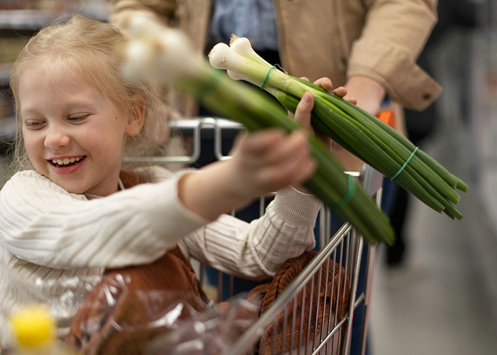 Happy child holding green onions in a shopping cart with an adult, capturing funny and inappropriate kids' moments shared by parents.