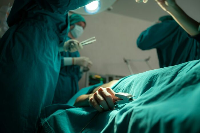 Patient hand gripping pulse oximeter in a dimly lit operating room during a tense medical procedure.