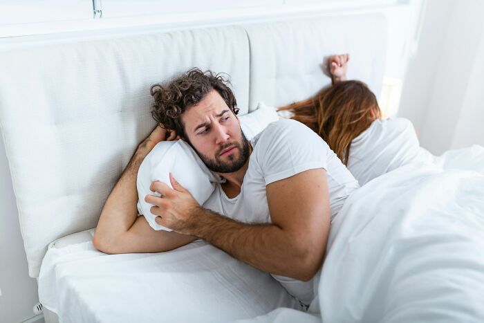 A man looking upset while lying in bed next to a woman with back turned, reflecting on driving away potential life partners.