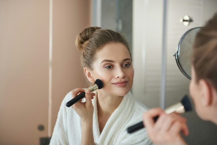 Young woman in a white robe applying makeup with a brush, reflecting calm despite no peaceful solution to our current situation.
