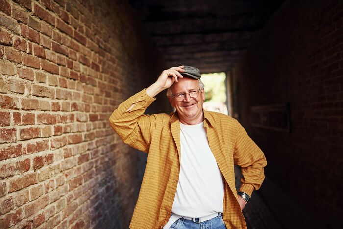 Smiling older man in a casual yellow shirt and cap standing in a brick alley, reflecting on personal mysteries and human nature.