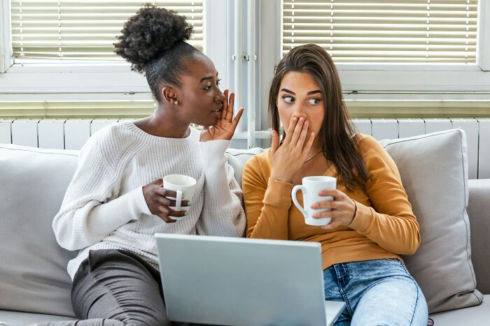 Two women sitting on a couch with coffee cups, one whispering to the other, showing moments realizing a close friend is a jerk.