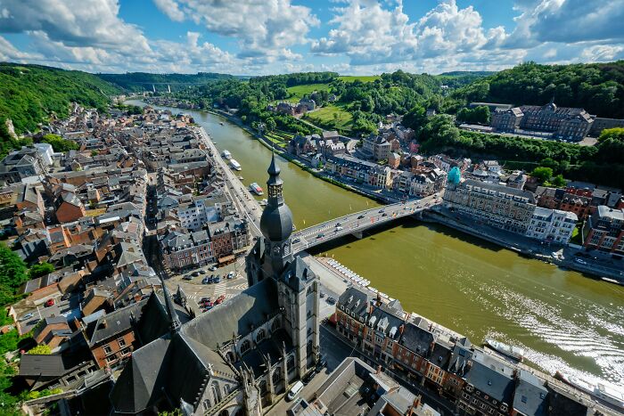 Aerial view of a historic European town by a river under a bright blue sky with clouds and green hills.