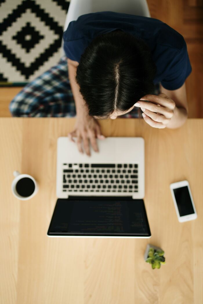 Person experiencing scary online moments, looking stressed while using a laptop at a wooden desk with coffee and phone nearby