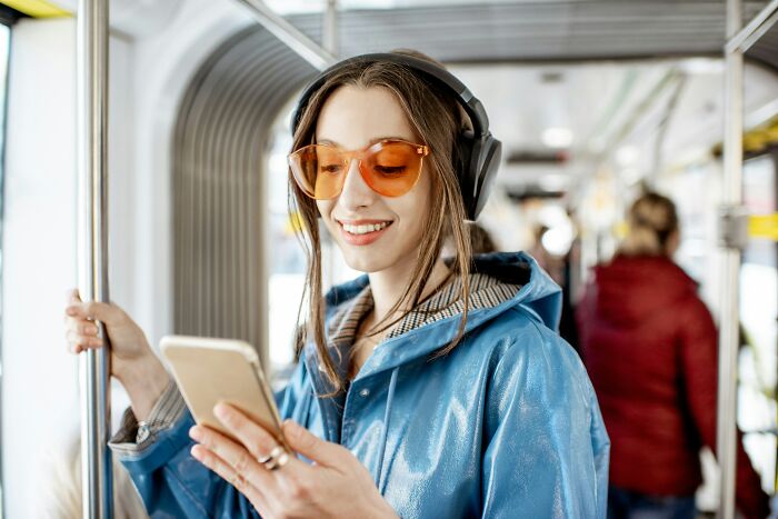 Young woman wearing headphones and orange glasses, smiling while using smartphone on public transport, representing legal total psychopath lifestyle.