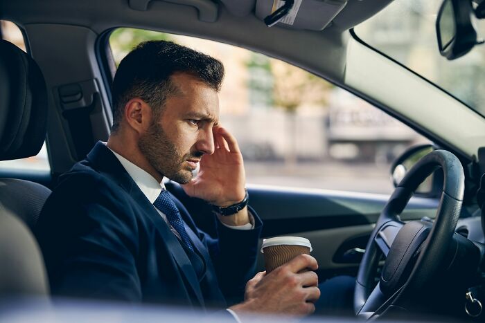 Man in a suit sitting in car holding coffee, looking frustrated and stressed, reflecting on unhinged ways to get back at cheating exes