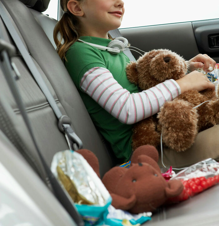 Young girl holding a teddy bear in a car surrounded by snacks, reflecting on stories about the worst parents seen.