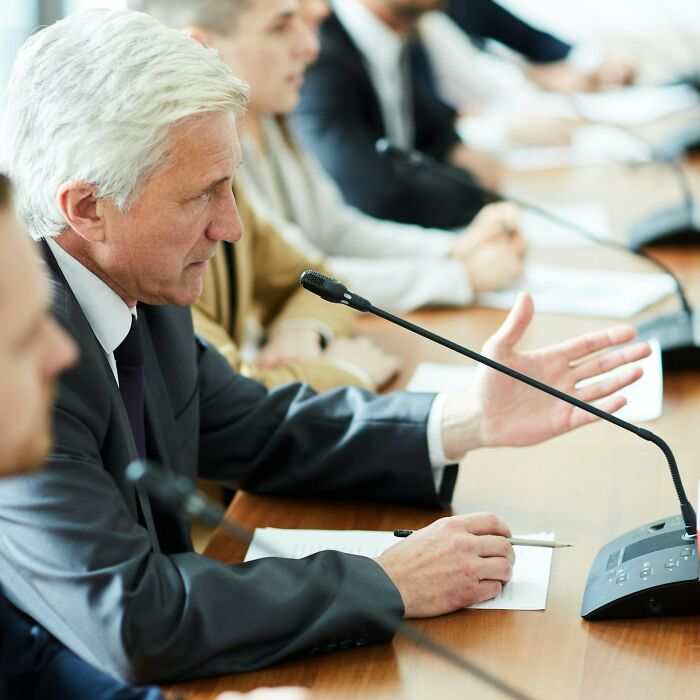 Man in suit speaking into microphone during a formal meeting where people share their wokest opinions on current situation.