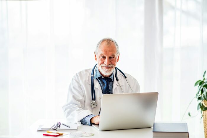 Older male doctor with stethoscope using laptop in bright office, symbolizing frustrating experiences with dismissive doctors.
