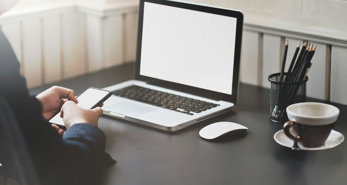 Person holding smartphone near open laptop with blank screen on desk, illustrating scary online moments and trauma.