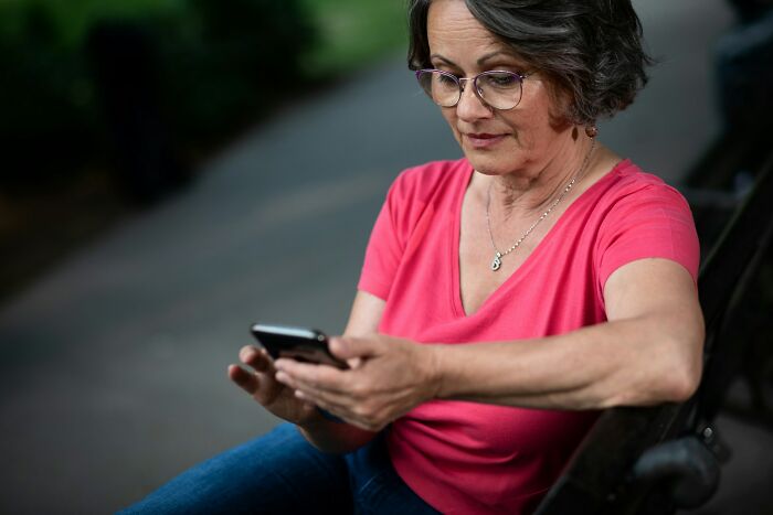 Woman in glasses and pink shirt sitting outdoors on a bench, focused on her phone, showing weird things couples started doing.